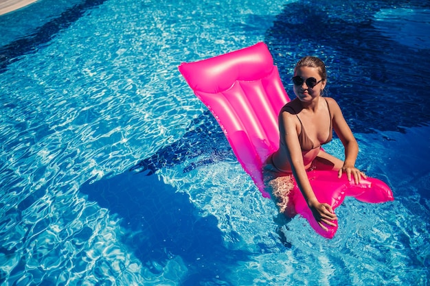 Sexy vrouw in zonnebril rusten en zonnebaden op een roze matras in het zwembad jonge vrouw in beige bikini zwembroek drijvend op opblaasbare roze matras