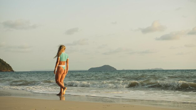 Sexy vrouw lopen alleen op strand kustlijn en ontspannen warme gouden zonsondergang vrouw lopen op water in blauwe zwembroek en zonnebril Concept rust tropische resort reizen toerisme gelukkig zomervakantie