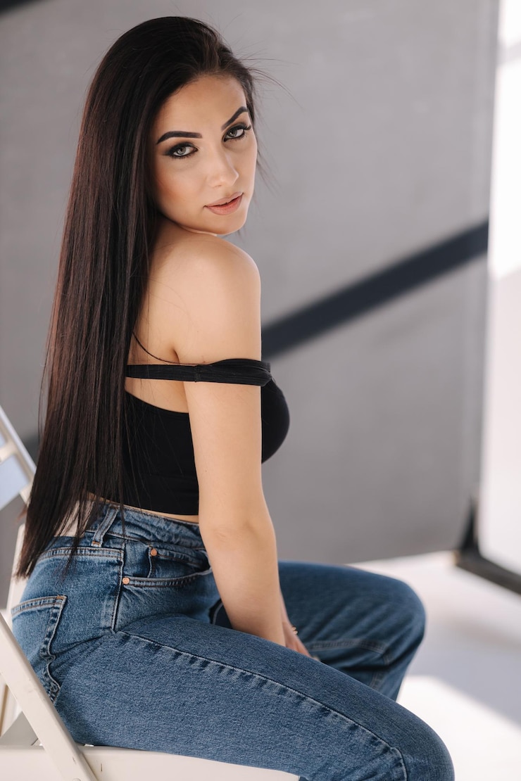 Photo sexy woman in black topic and blue jeans sits on chair during studio photoshoots brunette girl