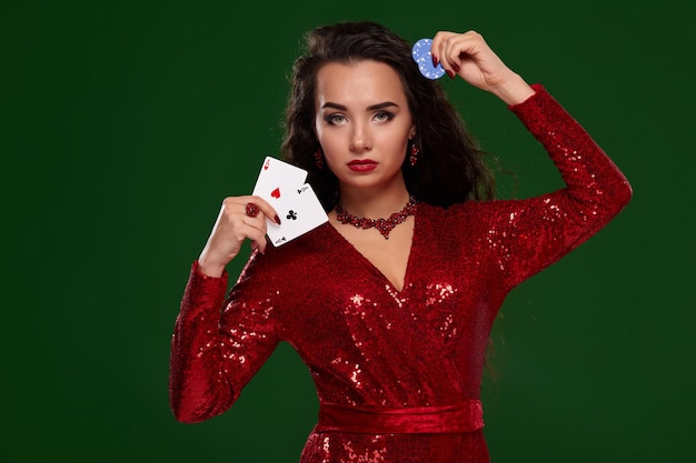 완벽한 헤어스타일과 이브닝 메이크업이 있는 빨간색 반짝이 드레스를 입은 섹시한 여자. 그녀는 녹색 배경에 진지해 보이는 카드 놀이와 도박 칩을 손에 들고 있습니다. 카지노