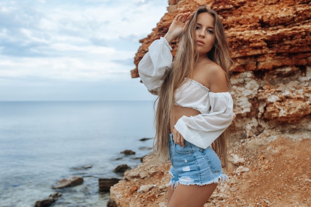 Sexy woman on a rocky beach in min shorts and a white\
blouse