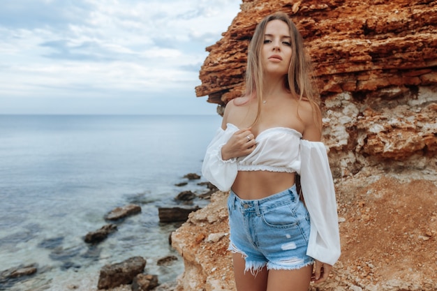 Sexy woman on a rocky beach in min shorts and a white\
blouse