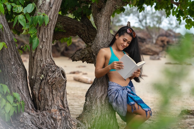 Donna sexy che sorride leggendo un libro sotto gli alberi sulla spiaggia