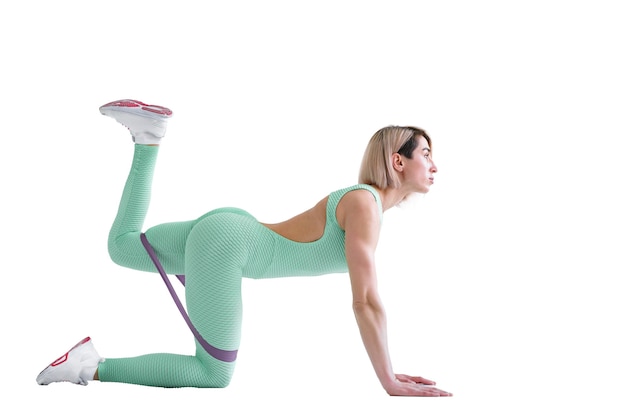 Sexy woman in sportswear using a resistance band in her exercise routine. Young woman performs fitness exercises on white wall. Isolate