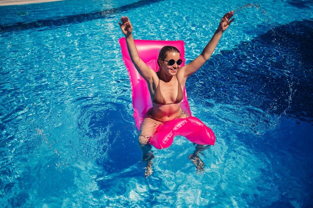 Sexy woman in sunglasses resting and sunbathing on a pink mattress in the pool Young woman in beige bikini swimsuit floating on inflatable pink mattress