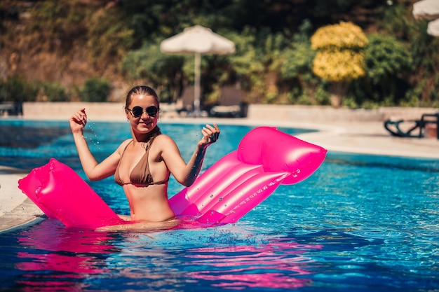 선글라스를 끼고 수영장에 있는 분홍색 매트리스에서 일광욕을 하는 섹시한 여성 풍선 핑크 매트리스에 떠 있는 베이지색 비키니 수영복을 입은 젊은 여성