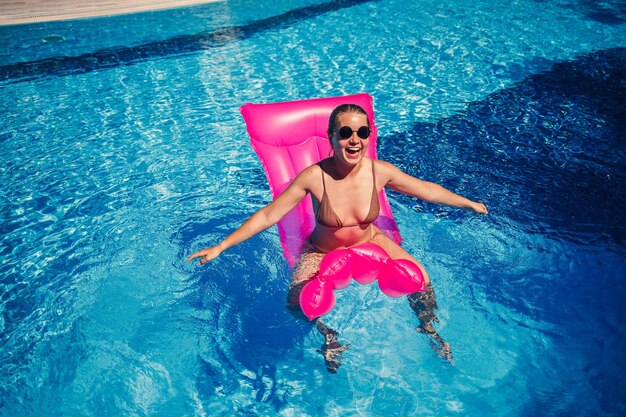 Sexy woman in sunglasses resting and sunbathing on a pink mattress in the pool Young woman in beige bikini swimsuit floating on inflatable pink mattress