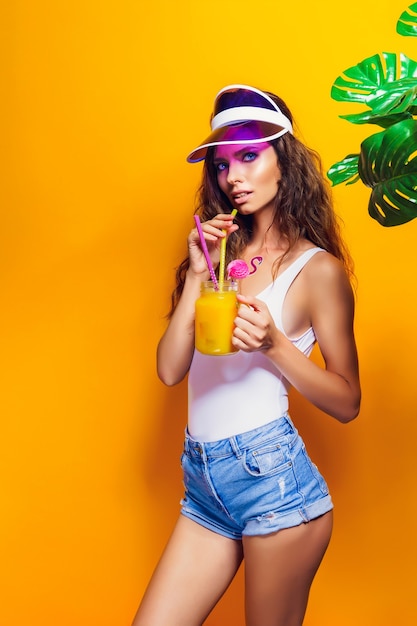 Sexy Woman in white swimsuit and blue jeans shorts, trendy visor holding glass of fresh drink while standing on yellow, orange hot