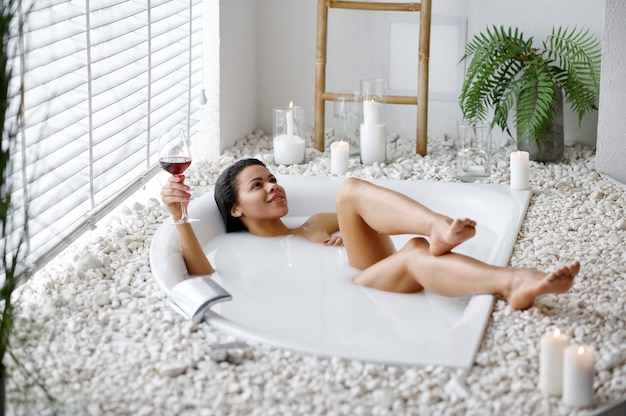 Sexy woman with glass of beverage in bath with milk. Female person in bathtub, beauty and health care in spa, wellness treathment in bathroom, pebbles and candles on background