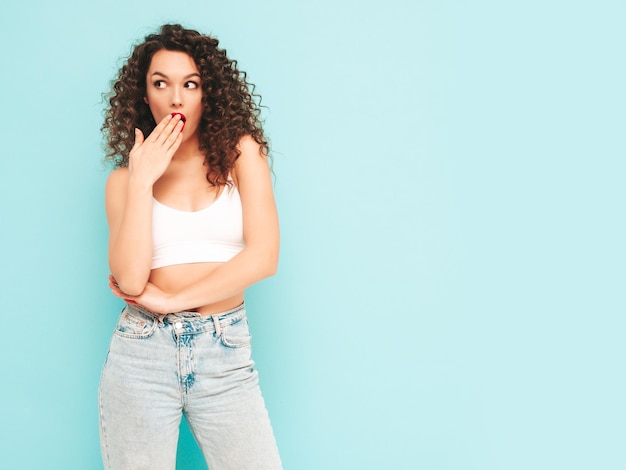 Sexy young beautiful smiling female in trendy summer hipster clothes carefree woman posing near blue wall in studio Positive brunette model with afro curls hairstyle Cheerful and happy