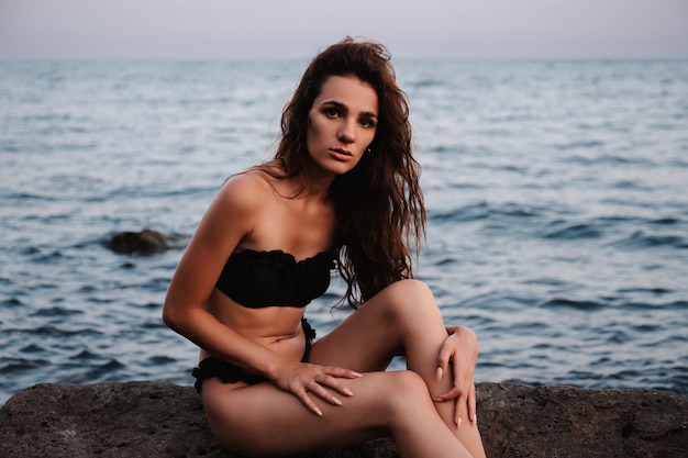 Sexy young girl in a black swimsuit sitting on the rocks by the sea in summer on vacation