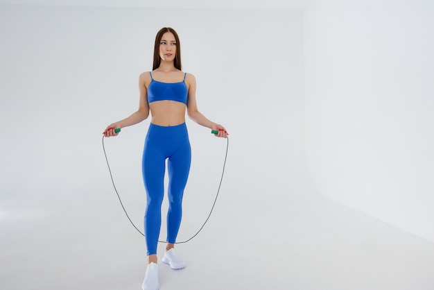 Sexy young girl jumping rope in a blue tracksuit on a white space. Fitness, healthy lifestyle.