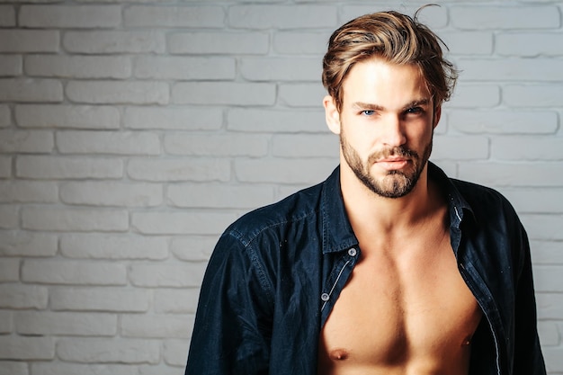 Sexy young handsome bearded man or guy with bare muscular chest in black shirt on white brick wall background copy space