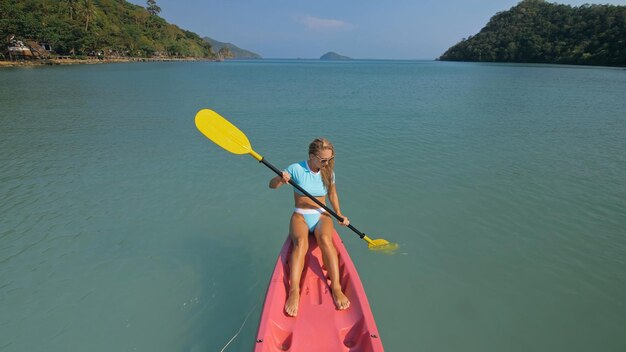 Sexy young woman in blue swimsuit swings feet in water on pink\
canoe on sea against hills backside