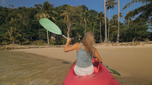 Sexy young woman in blue swimsuit swings feet in water on pink\
canoe on sea against hills backside