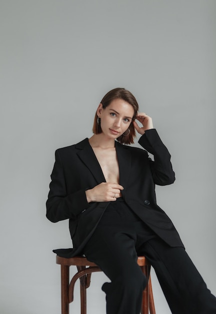 Sexy young woman in a jacket on a naked body and trousers sitting on chair in the studio Fashion shot