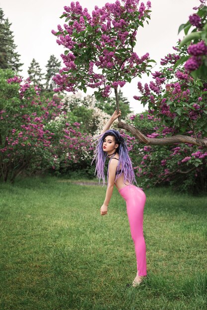 sexy young woman model with color dreadlocks, in pink leather pants is standing under the lilac