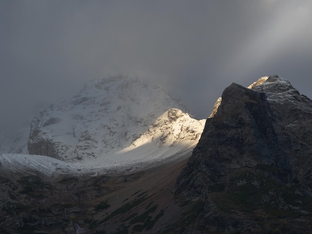 Sfeervol landschap met scherpe rotsen en hoge besneeuwde bergtop in sneeuwstorm lage wolken bij bewolking Dramatisch somber landschap met grote sneeuwbergen en gletsjer in grijze bewolkte lucht bij zonsopgang