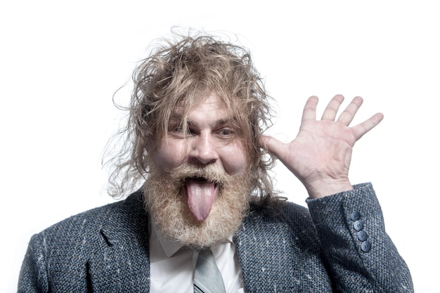 Shaggy adult grayhaired man with a beard wearing a gray suit shows tongue and holds a hand near the ear Isolated