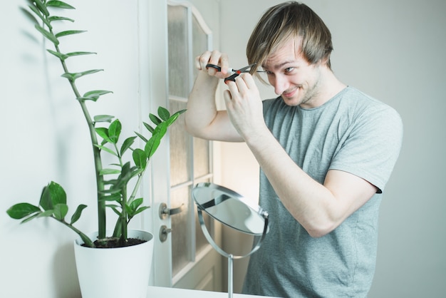 A shaggy man in a gray T-shirt with scissors in his hands is trying to cut his hair while looking in the mirror. Stay home. Quarantine.