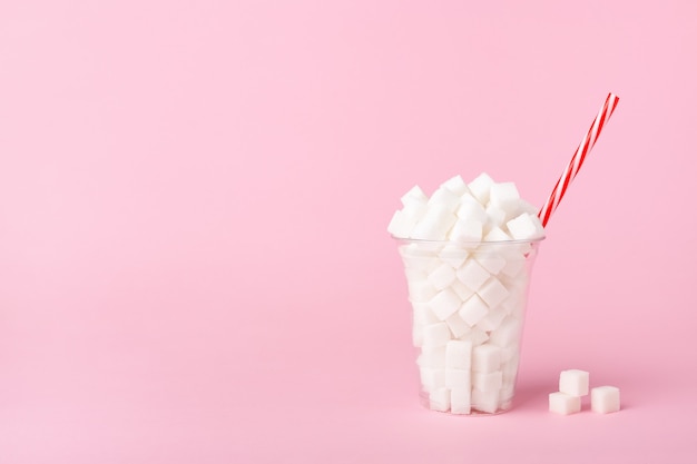 Shake glass full of sugar cubes on pastel pink background. Unhealthy food concept. Copy space, side view.