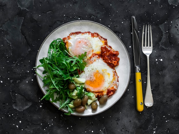 Shakshuka アボカド ルッコラ サラダ オリーブの朝食ランチ ディナー暗い背景のトップ ビューで