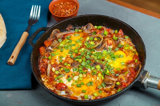 Shakshuka in een koekenpan op een grijze achtergrond naast groenten en brood op een blauw servet