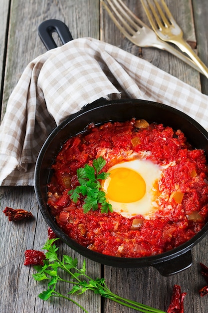 Shakshuka with tomato sauce and fried egg in a cast iron pan for breakfast on the old wooden table