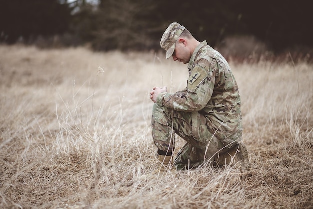 shallow focus shot of a young soldier praying while kneeling on a dry grass
