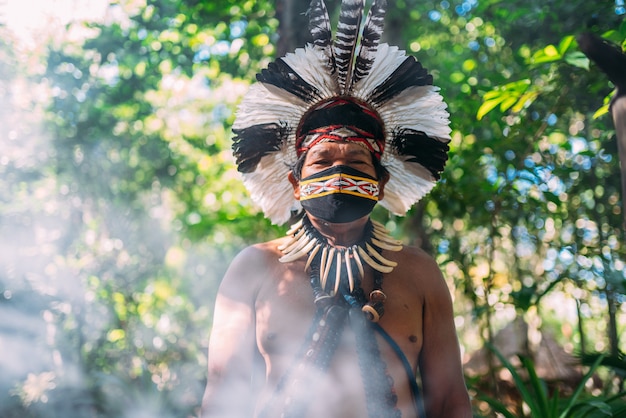 Shaman of the PataxÃ³ tribe. Elderly Indian man wearing feather headdress and face mask due to the covid-19 pandemic