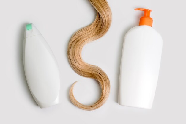 Shampoo and conditioner bottles and lock of curly blonde hair isolated on light background, top view. Flat lay, copy space for text. Hair care cosmetics, beauty haircare products, hair treatment.