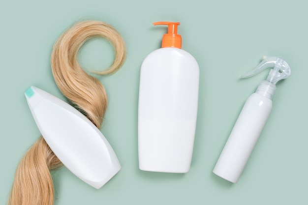Shampoo wrapped in lock of curly blonde hair, conditioner bottle and hair spray mockups on mint background, top view. Flat lay in pastel colors. Hair care cosmetics, haircare products, hair treatment.