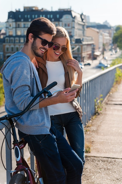 Sharing photos with friends. Side view of beautiful young couple looking at the mobile phone and smiling while standing near bicycle outdoors