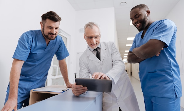 Sharing positive vibes among collective. Easygoing upbeat aged physician working in the hospital and using modern technical device while having conversation with colleagues