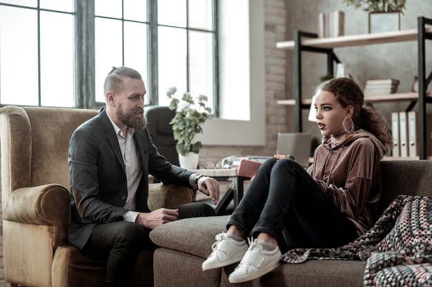 Sharing problems. Curly emotional teenager sitting on sofa and sharing her problems with counselor