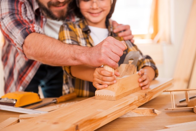 Sharing skills with his son. Close-up of smiling young male carpenter teaching his son to work with wood in his workshop