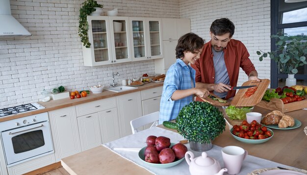 スキルを共有する若いお父さんと彼の小さな幸せな息子が一緒に野菜サラダを準備する