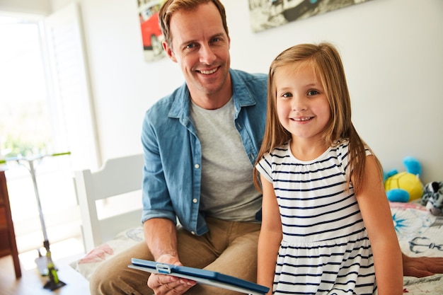 父親とオンラインで時間を共有するデジタルタブレットを使用して自宅のリビングルームに一緒に座っている父親と彼の若い娘のショット