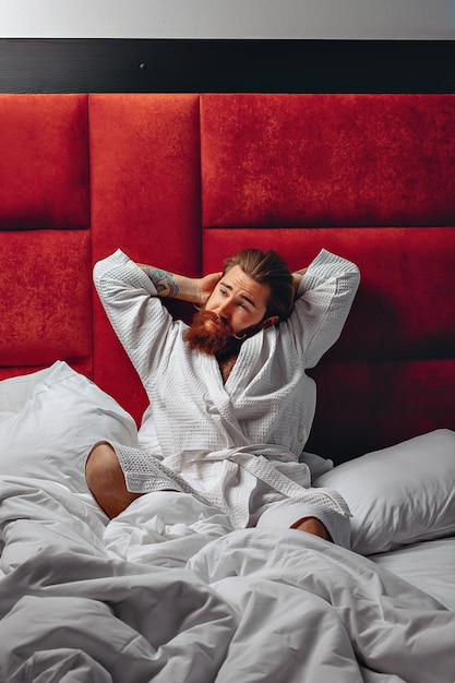 Sharming pretty adult man just awakened man in a robe with a long stylish mustache and beard sitting on the bed and stretching his arms up. Awake concept