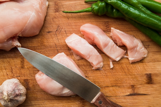 Photo sharp knife and raw chicken slices on cutting board with garlic and green chili