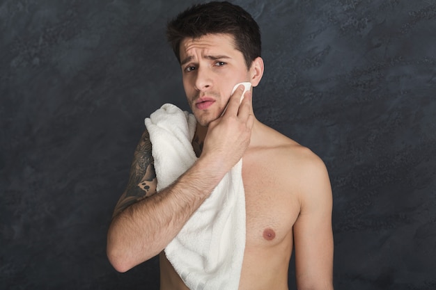 To shave or not to shave. Handsome pensive tattooed man touching his face while standing with towel on shoulders, gray studio background