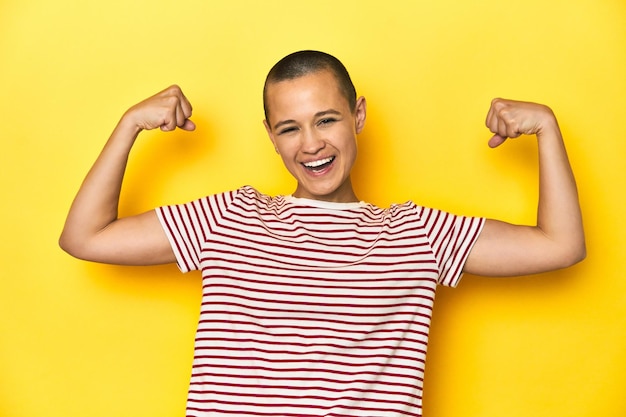 Photo shaved head woman in red striped tee yellow backdrop showing strength gesture with arms symbol