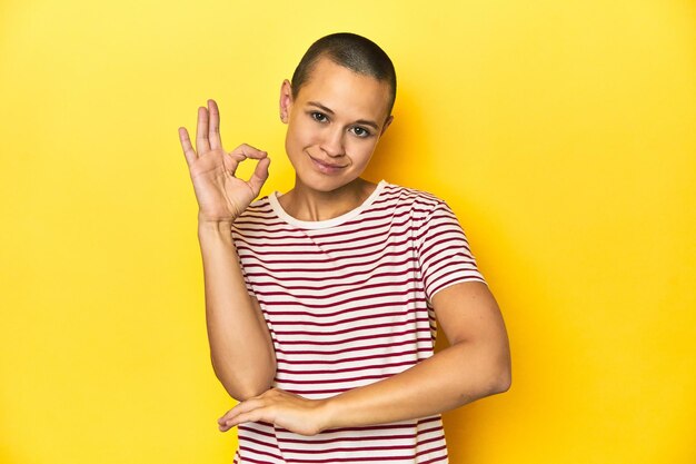 Photo shaved head woman in red striped tee yellow backdrop winks an eye and holds an okay gesture