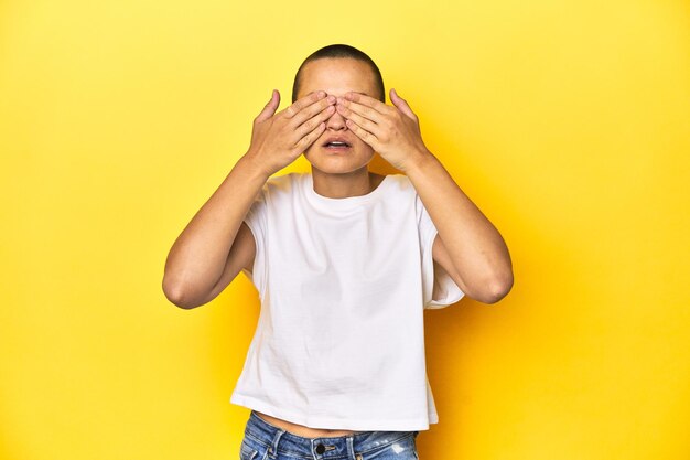 Shaved head woman in white tank top yellow backdrop afraid covering eyes with hands