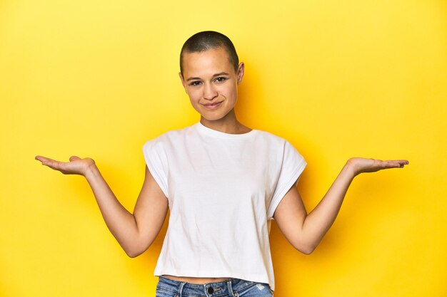 Shaved head woman in white tank top yellow backdrop makes scale with arms feels happy and confident