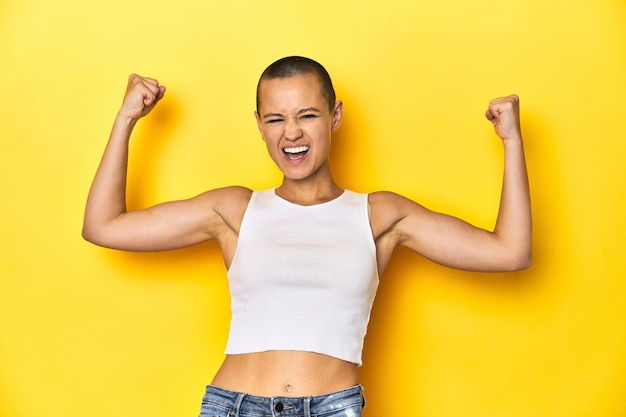 Shaved head woman in white tank top yellow backdrop raising fist after a victory winner concept