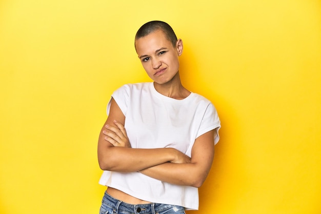 Shaved head woman in white tank top yellow backdrop unhappy looking in camera