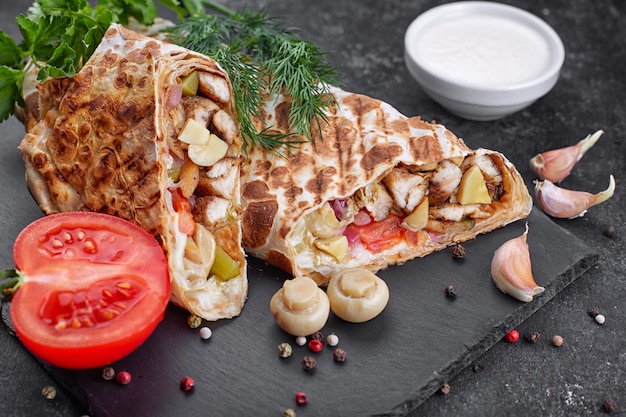 Shawarma with chicken meat, with sauce, onions, pickles, tomato, garlic, herbs and mushrooms champignons, on slate, against a dark concrete background