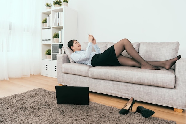 she is playing on her smartphone and also working at home by using notebook online at the same time lying down on cozy sofa