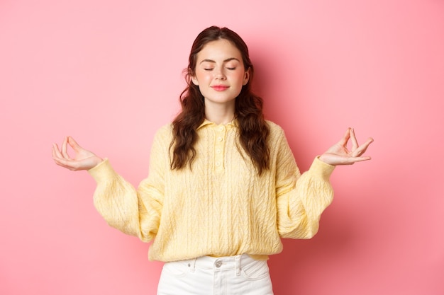 She is relaxed smiling young peaceful girl meditating holding hands in zen gesture close eyes feel relieved and calm standing against pink wall